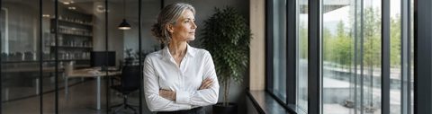 Transformations-Coaching für Change-Leader: Frau in Business-Dress steht im Büro und blickt aus dem Fenster